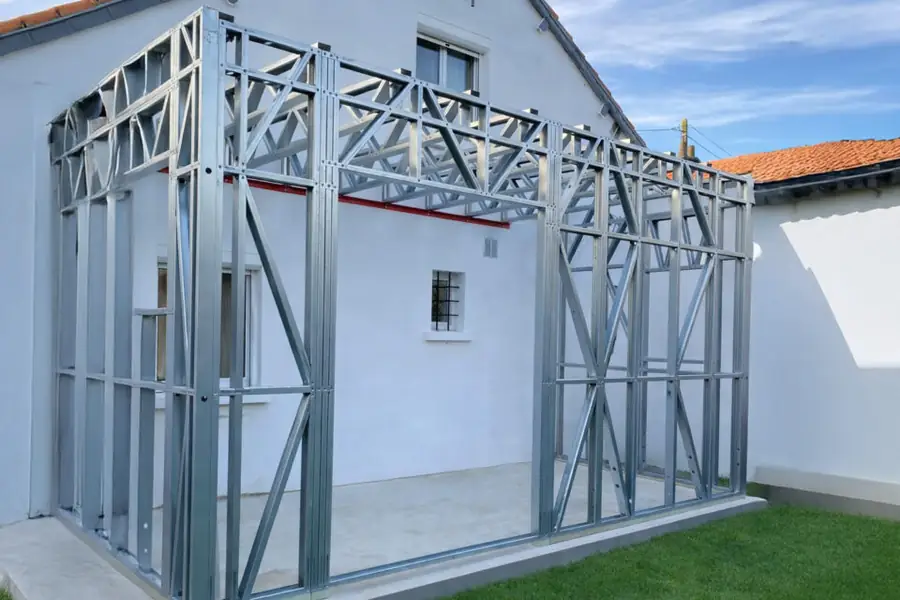 Ossature métallique d'une extension accolée au mur enduit d'une maison existante, jonction entre le neuf et l'ancien visible.