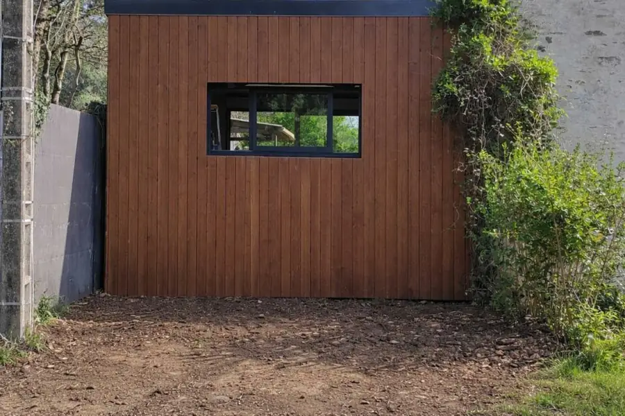 Extension terminée en bardage bois vertical à toit plat, greffée sur le pignon d'une maison en pierre.