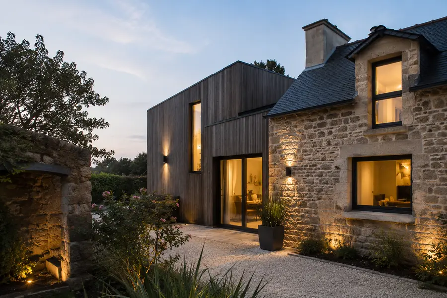 Maison en pierre surélevée d'un niveau en bois, pièces éclairées à la tombée de la nuit.