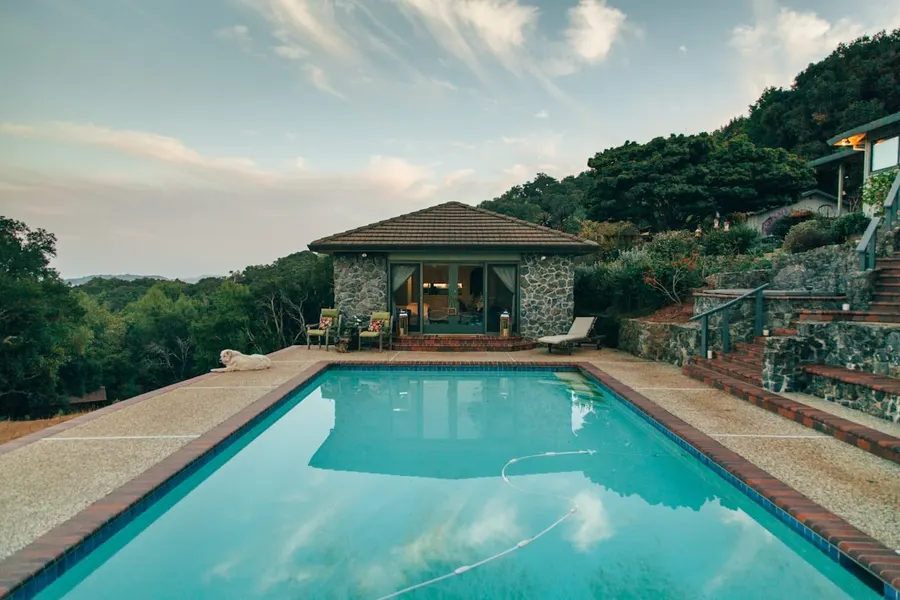 Piscine enterrée rectangulaire devant un pool house en pierre, sur un terrain en pente soutenu par des murets et des escaliers.